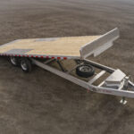 Two-axle aluminum flatbed trailer with a wooden deck, spare tire mounted on the frame, and a raised rear ramp on a dirt surface.