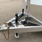 Close-up of a utility trailer tongue showing the trailer jack, hitch coupler, safety chains, and diamond-plate fender on asphalt surface (informative image).