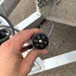 Hand holding a circular 7-pin trailer electrical connector with metal pins in a black housing.