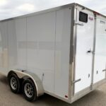 White enclosed cargo trailer with dual axles and rear double doors in a parking lot area.