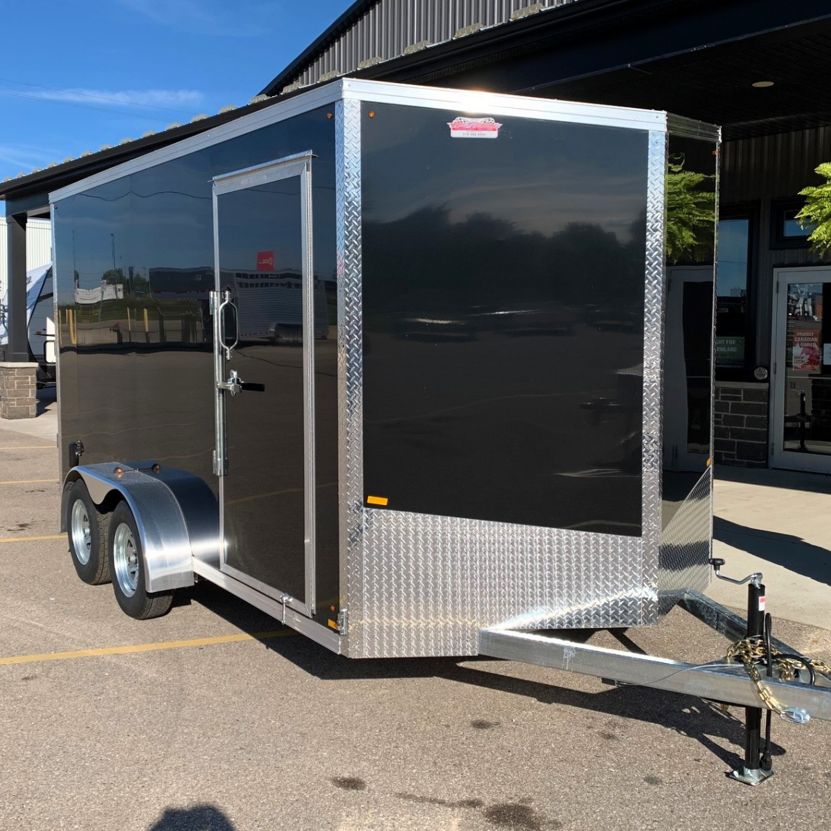 Black enclosed cargo trailer with diamond-plate trim and tandem wheels, parked outside a building.