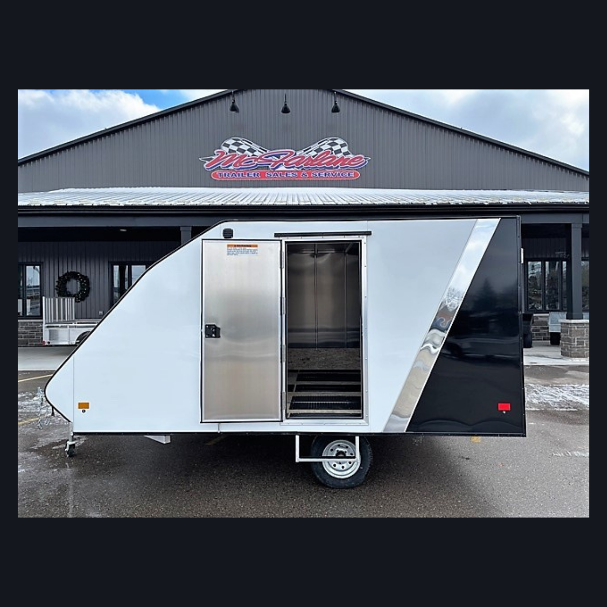 White and black travel trailer parked in a lot with its door open and steps visible, in front of a showroom building with a sign above the entrance.
