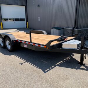 Flatbed trailer with wooden deck and dual axles parked in front of a gray industrial building and a large white garage door.