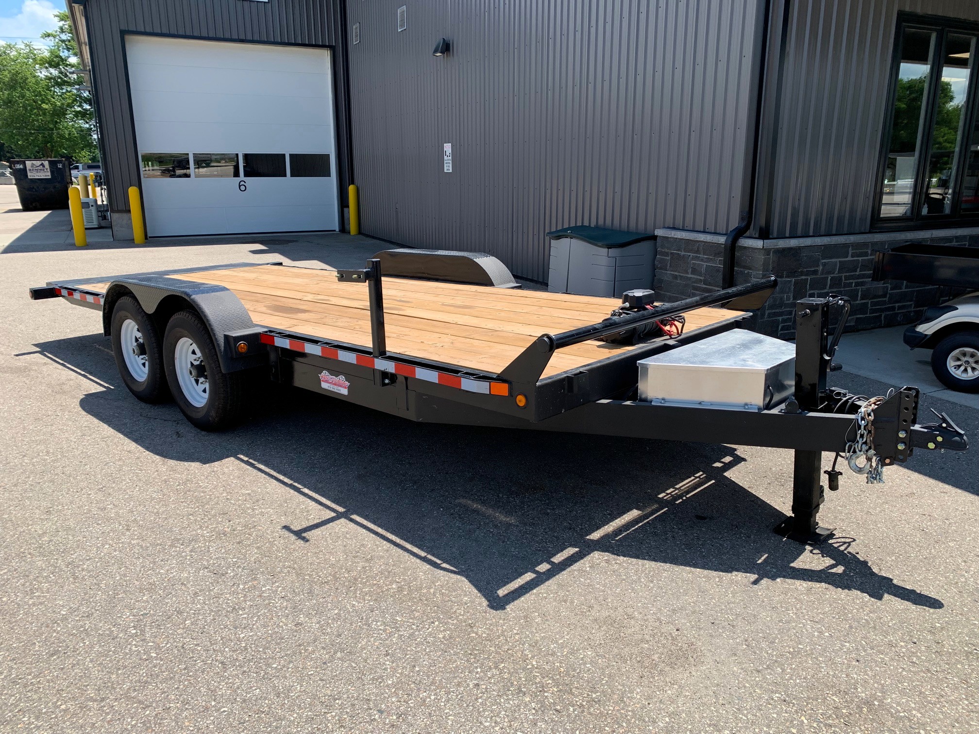 image0 52 - McFarlane Trailer Sales Flatbed trailer with wooden deck and dual axles parked in front of a gray industrial building and a large white garage door.