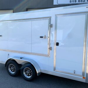 White enclosed cargo trailer with two side doors and chrome trim, parked near a building; rainbow reflection visible on its side near the top edge.