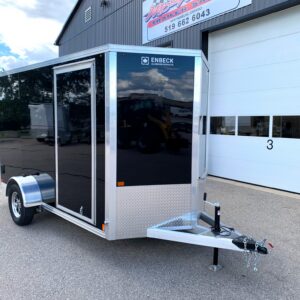 Front corner view of a black enclosed trailer with aluminum trim and diamond-plate front, parked in a lot near a building sign.