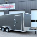 Gray enclosed cargo trailer with diamond-plate front and tandem wheels, hitched in a parking lot in front of a commercial building with a trailer sales sign