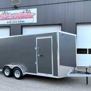 Gray enclosed cargo trailer with diamond-plate front and tandem wheels, hitched in a parking lot in front of a commercial building with a trailer sales sign