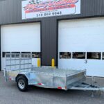 Silver aluminum utility trailer with a mesh gate and long hitch, parked in front of a dark corrugated metal building with large white garage doors.