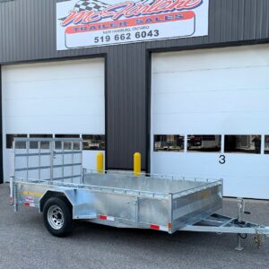 Silver aluminum utility trailer with a mesh gate and long hitch, parked in front of a dark corrugated metal building with large white garage doors.