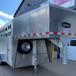 Silver livestock trailer with a spare tire mounted on the side, standing on its support legs beside a building with red-and-white reflective stripes along the bottom edge of the trailer side restricted.