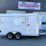 White enclosed cargo trailer with two rear doors and chrome trim, parked in front of a grey industrial building.