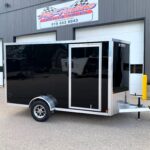 image1 54 - McFarlane Trailer Sales Black enclosed cargo trailer with chrome trim and diamond plate front, parked outside a metal industrial building with a sign above.