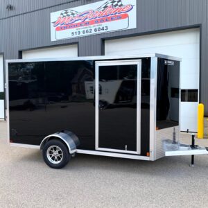 Black enclosed cargo trailer with chrome trim and diamond plate front, parked outside a metal industrial building with a sign above.