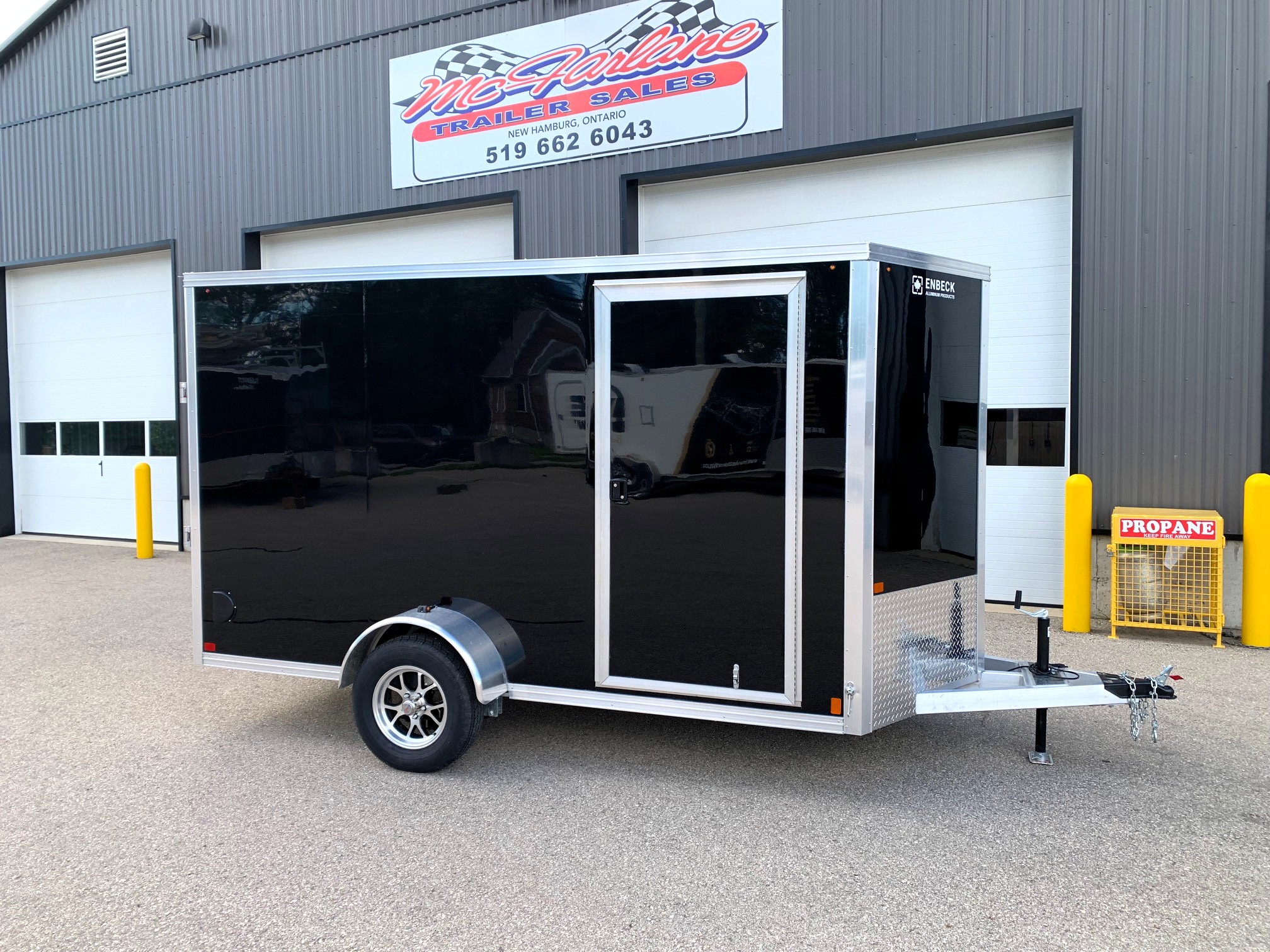 image1 54 - McFarlane Trailer Sales Black enclosed cargo trailer with chrome trim and diamond plate front, parked outside a metal industrial building with a sign above.