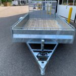 Metal utility trailer with a wooden deck and diamond-mesh gates at a parking lot near a building, ready to hitch up.