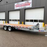 Flatbed trailer parked in front of a gray industrial building with three large bay doors and yellow safety bollards nearby.