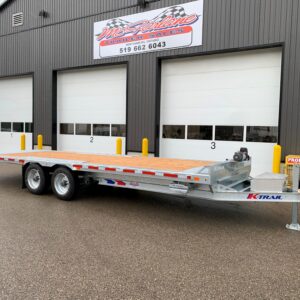 Flatbed trailer parked in front of a gray industrial building with three large bay doors and yellow safety bollards nearby.