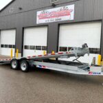 Tilt-bed trailer with fold-down ramps parked in front of a gray industrial building; sign reads McFarlane Trailer Sales.
