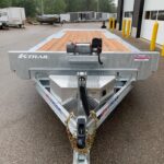 Front view of a silver K-Trail flatbed trailer with a wooden deck and winch, parked among other trailers in an industrial lot.