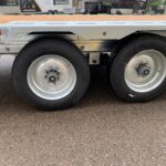 Side view of a flatbed trailer with two large wheels, silver rims, and diamond‑plate fender; background shows other trailers and equipment.