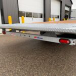 Diamond-plate loading ramp on a flatbed trailer at an industrial bay with yellow bollards in the background, warehouse doors visible behind it.