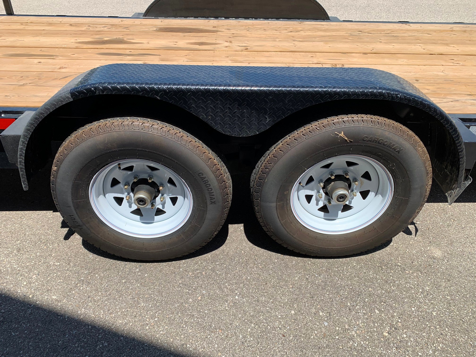 image2 48 - McFarlane Trailer Sales Dual-axle trailer with diamond-plate fenders over two tires and a wooden bed visible above the wheels, on a paved surface.