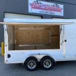 White dual-axle trailer with a large side opening, parked outside a gray metal industrial building with a sign above.
