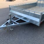 Galvanized utility trailer with a wooden deck and metal rails on a paved lot, front hitch and safety chains visible