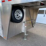 image2 55 - McFarlane Trailer Sales Trailer side with a mounted spare tire inside a wheel well and a stabilizing leg extended to the ground on a paved surface, aluminum siding visible above.