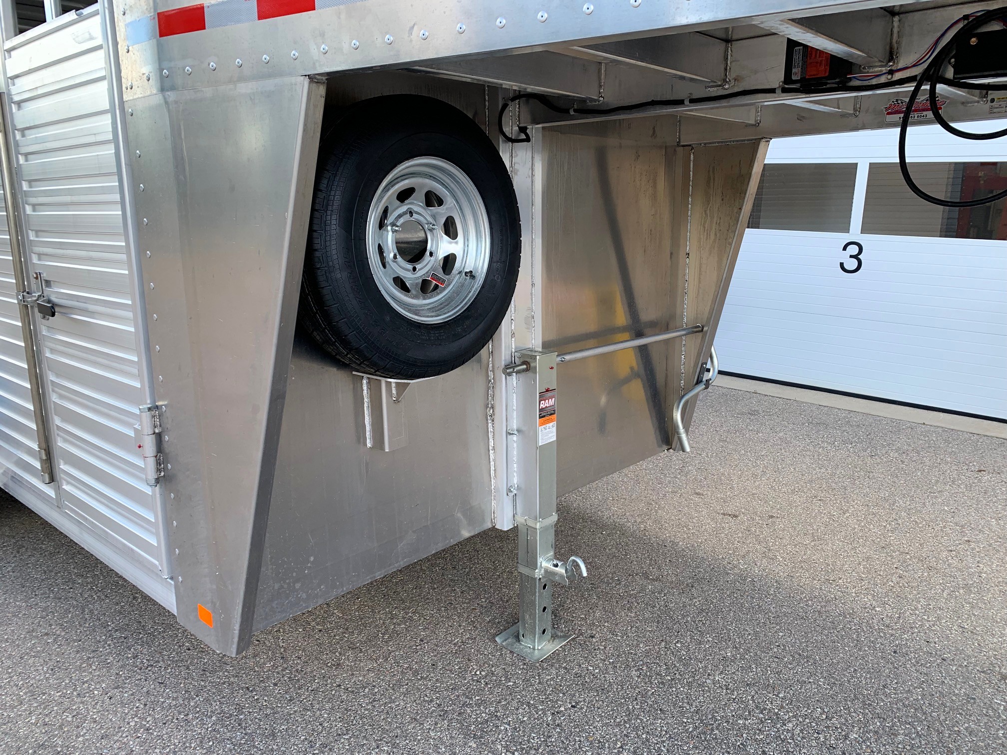 image2 55 - McFarlane Trailer Sales Trailer side with a mounted spare tire inside a wheel well and a stabilizing leg extended to the ground on a paved surface, aluminum siding visible above.