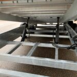 Underside view of a metal trailer frame showing hydraulic suspension, beams, and wheels in the background.