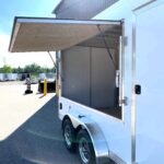 Rear of a white enclosed trailer with its ramp-style door propped open, exposing an empty interior; parked in an outdoor lot with other trailers nearby