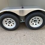 Close-up of a tandem trailer wheel setup with a curved silver fender and a small mounting bracket above the wheel.