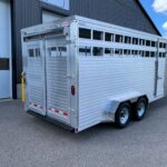 image3 50 - McFarlane Trailer Sales Silver aluminum livestock trailer with vented side slats tied to a dual-axle chassis, parked beside a dark industrial building on a sunny day
