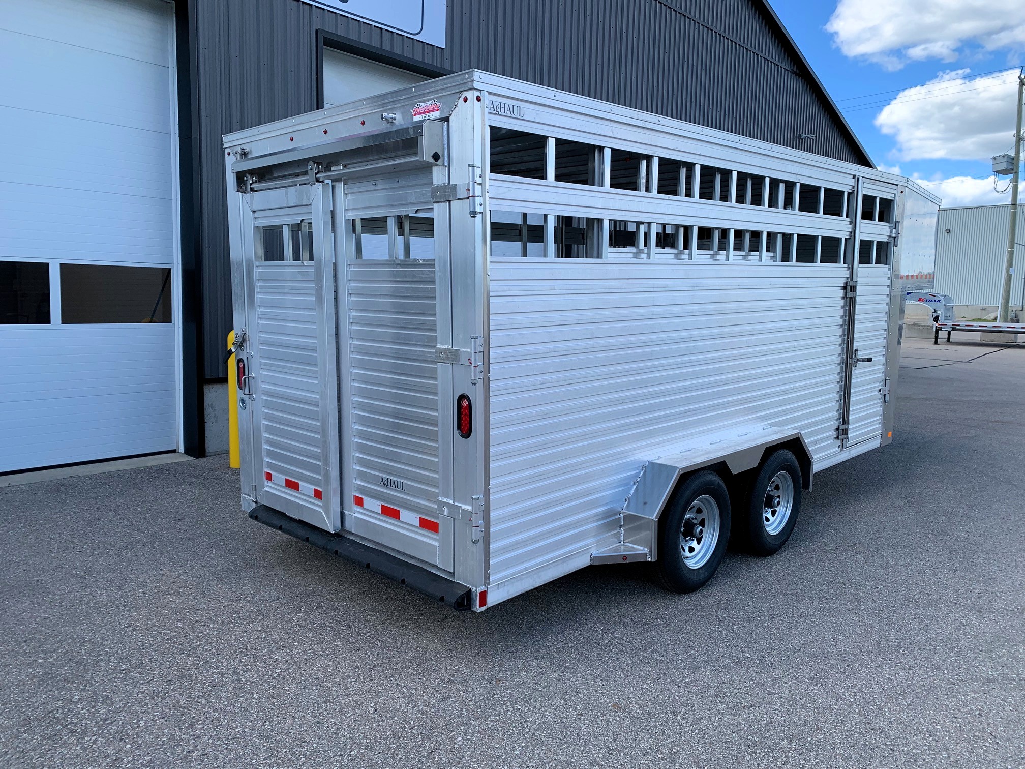 image3 50 - McFarlane Trailer Sales Silver aluminum livestock trailer with vented side slats tied to a dual-axle chassis, parked beside a dark industrial building on a sunny day