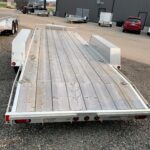 Metal flatbed trailer with wooden planks and diamond-plate fenders parked in an industrial lot, empty load area ready for use.