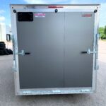 Rear of a silver double-door trailer with diamond-plate trim and latching mechanisms, outdoors under a blue sky (trailer brand stickers visible).