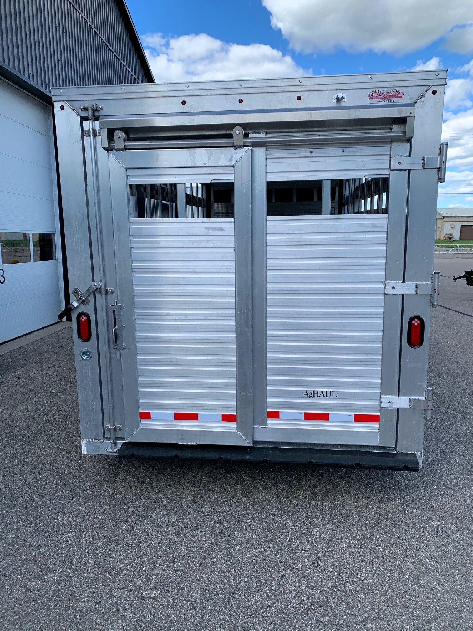 image4 49 - McFarlane Trailer Sales Rear of a silver metal livestock trailer with sliding doors and red reflectors, labeled 'AgHAUL' under a blue sky.