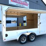 Open utility trailer with a large service window and plywood interior, parked in front of a metal building with a sign above indicating a trailer sales business