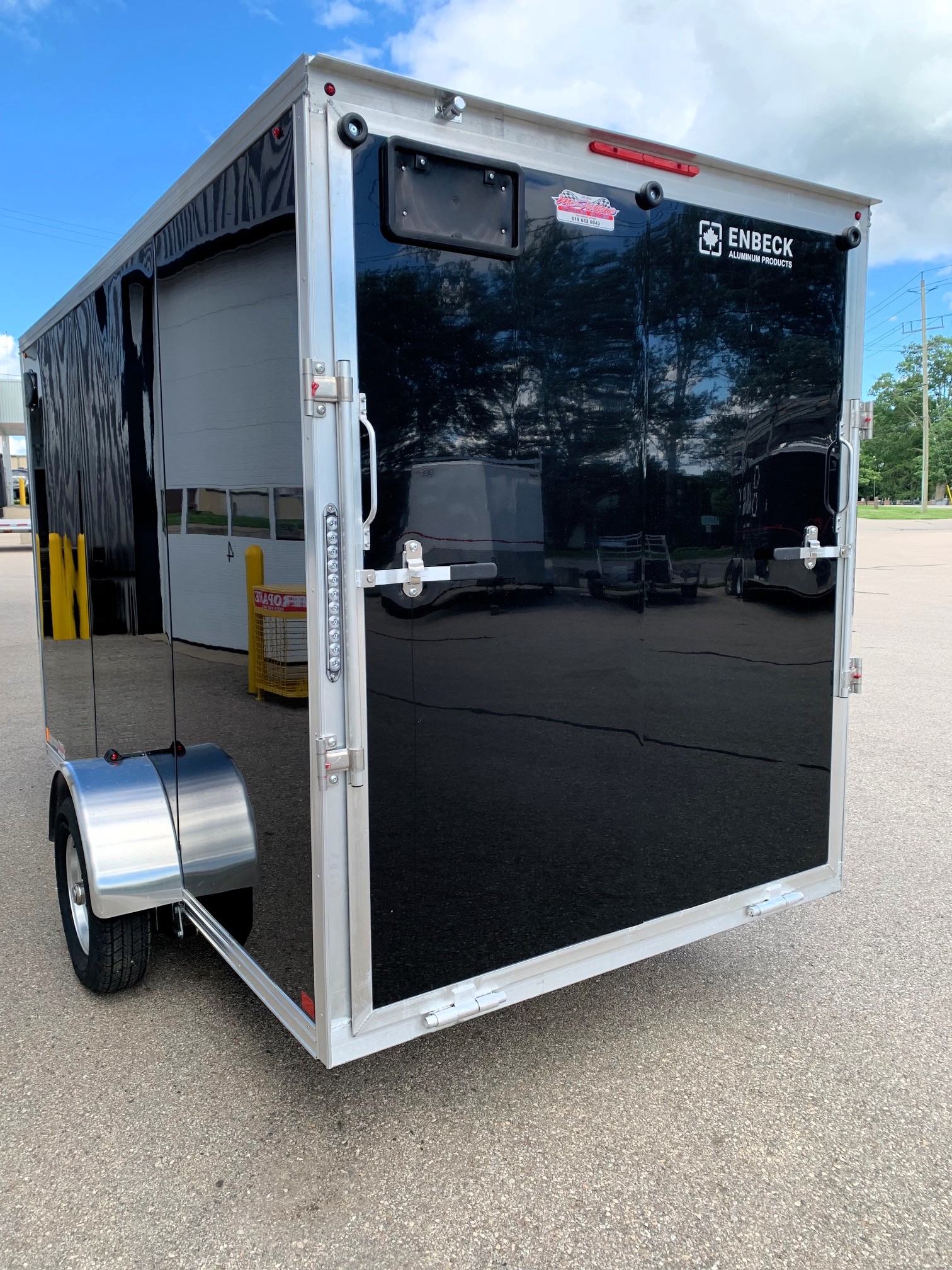 image5 38 - McFarlane Trailer Sales Black enclosed cargo trailer rear with 'ENBECK' logo and aluminum trim; open back doors with latches, wheel fender visible, outdoor lot two-door hatch reflections in glossy surface.