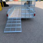 Metal utility trailer with a mesh gate and wooden bed in a parking lot, ramp lowered for loading.