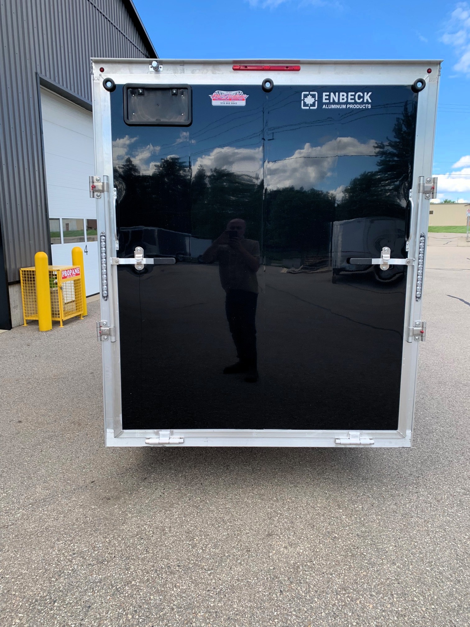 image6 28 - McFarlane Trailer Sales Rear view of a black enclosed trailer with silver frame and ENBECK Aluminum Products logo, reflecting a blue sky and clouds.