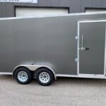Long gray enclosed trailer with diamond-plate trim, side access door, and tandem wheels parked on asphalt outside a building.