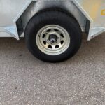 Close-up of a trailer wheel with a black tire and silver rim, mounted on a diamond-plate metal fender on a utility trailer.