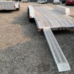 Aluminum flatbed trailer with diamond-plate ramps in a gravel lot, awaiting loading near a dark building.