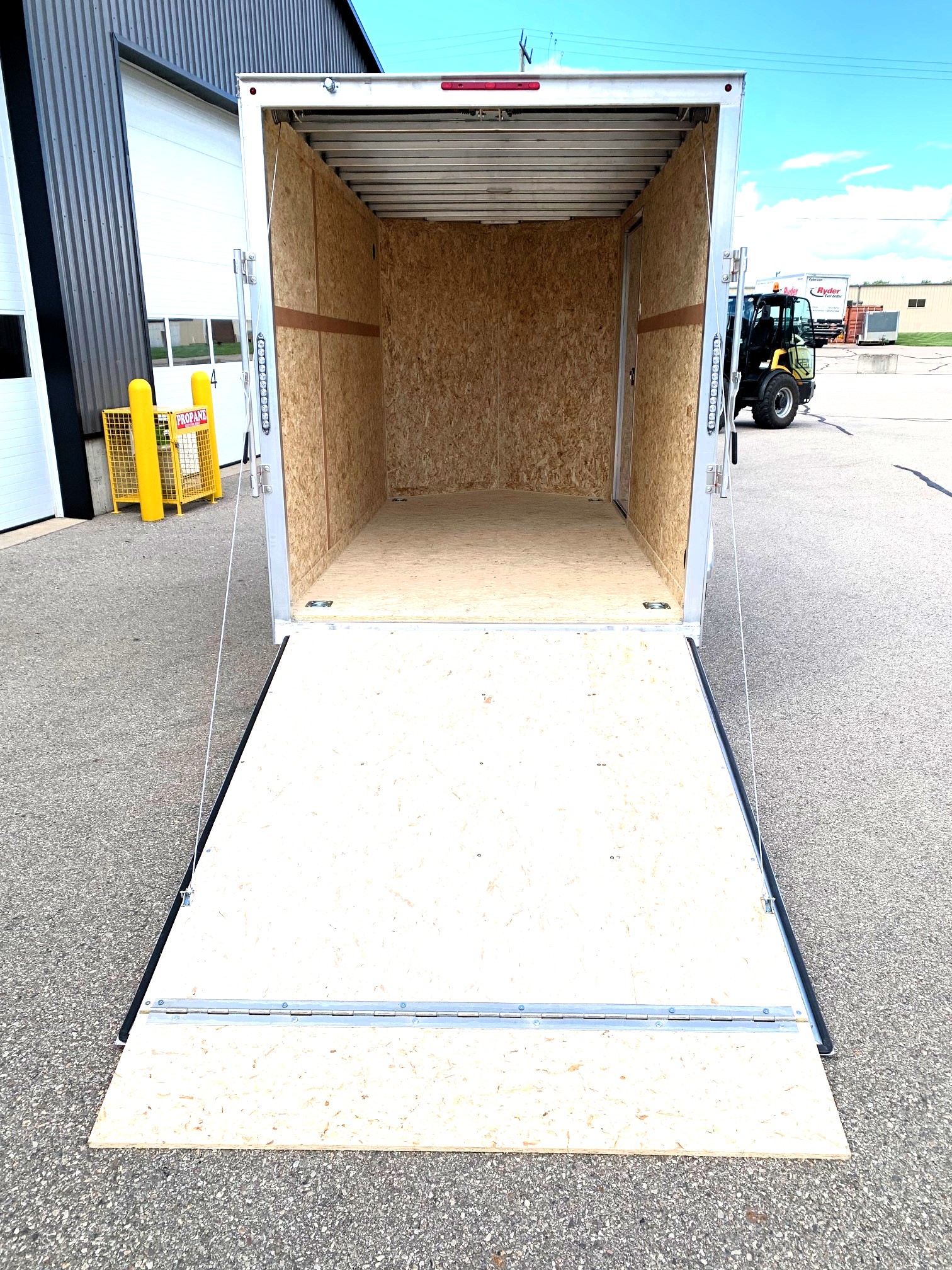 image7 24 - McFarlane Trailer Sales Open aluminum cargo trailer with plywood interior and lowered ramp, parked on asphalt outside an industrial building.