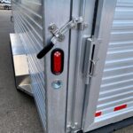image7 25 - McFarlane Trailer Sales Rear corner of a metal cargo trailer showing a latch bar, hinge, and red taillight on corrugated aluminum siding.