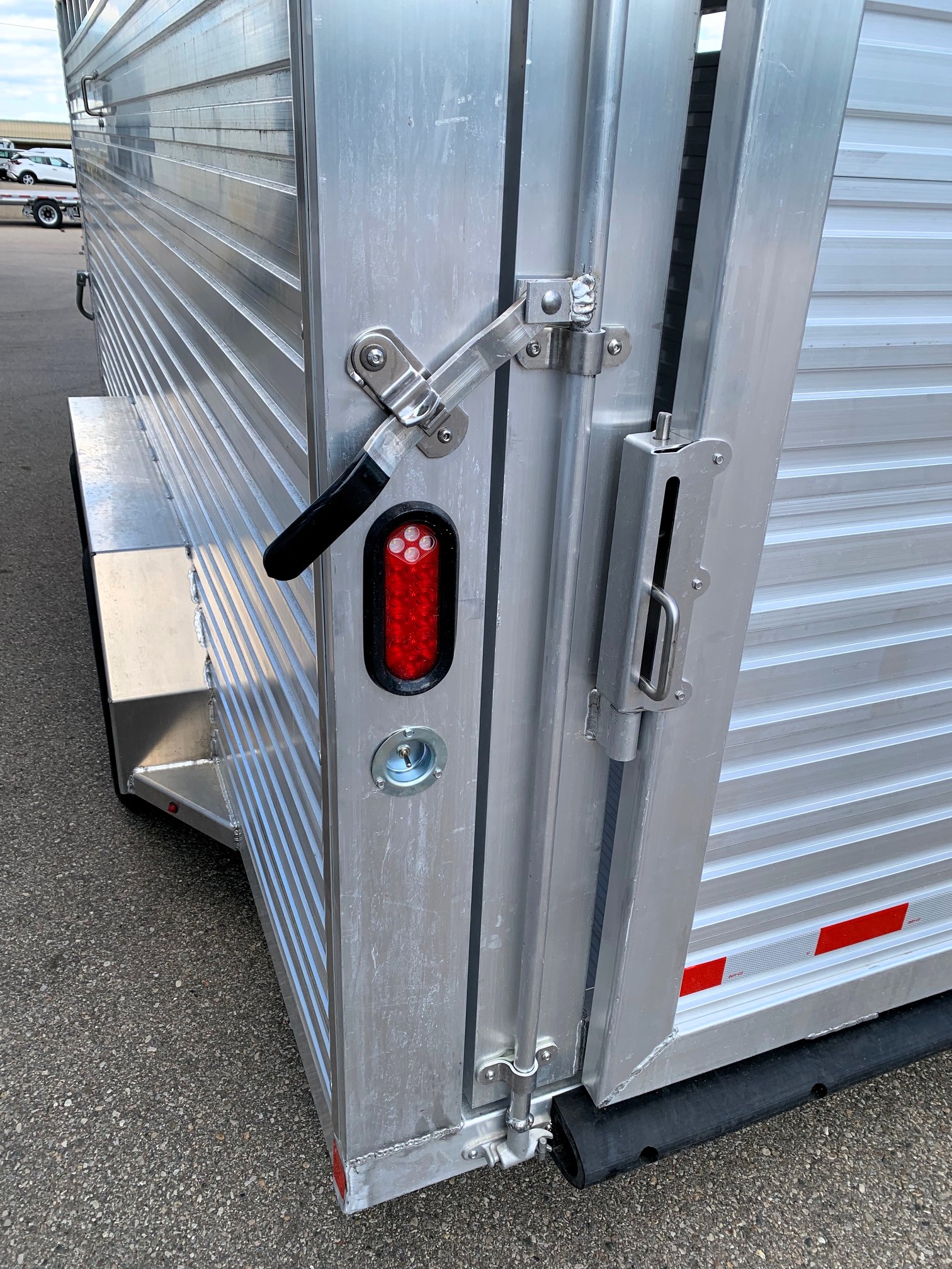 image7 25 - McFarlane Trailer Sales Rear corner of a metal cargo trailer showing a latch bar, hinge, and red taillight on corrugated aluminum siding.