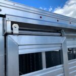 image8 15 - McFarlane Trailer Sales Close-up of a silver aluminum trailer side with a hinge, latch, and small red marker lights under a blue sky.